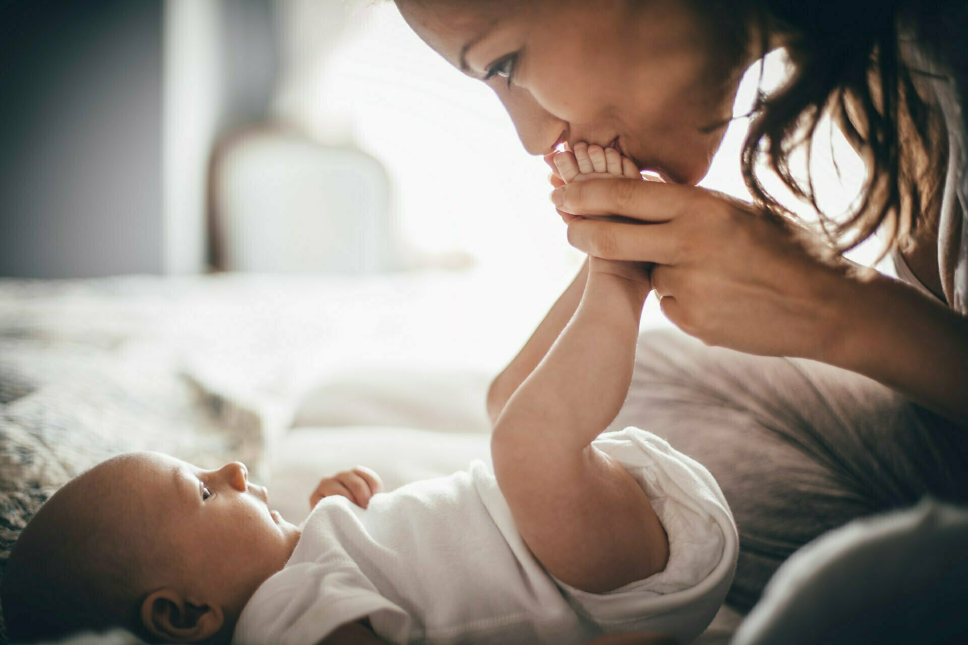 A happy mother kisses her baby's feet while she is lying down. The baby is looking up at her wearing a white baby grow.